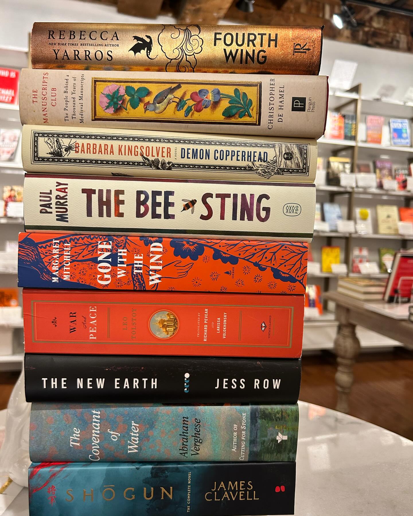 A stack of large books displayed on a table at the store: Fourth Wing, The Manuscripts Club, Demon Copperhead, The Bee Sting, Gone With the Wind, War and Peace, The New Earth, The Covenant of Winter, Shogun